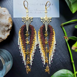 May include: Gold-tone earrings with a cross-shaped top and beaded fringe in shades of burgundy, black, yellow, and white. Dangling gold stars add detail. The earrings are handmade and feature the text "THE MOONLIT DREAMER".