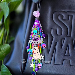 May include: A colorful beaded charm with an alien theme. The charm features a pink flower, a silver UFO, alien heads, and stars. The charm is attached to a black bag. The charm has the text "The Moon Dreamer".