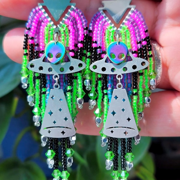 May include: A pair of handmade alien-themed earrings. The earrings feature a silver UFO and alien head charm, with a beaded design in shades of pink, green, black, and silver. The earrings are designed with a dangling, layered effect.