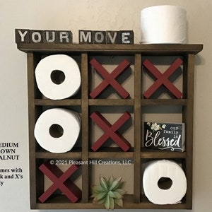 May include: A medium brown walnut tic-tac-toe-themed bathroom shelf with red X's and white O's. The shelf holds toilet paper rolls and a small sign that says "Our family is Blessed". The top shelf has the words "YOUR MOVE".