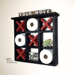 May include: A black wooden wall-mounted shelf with a tic-tac-toe design. The shelf holds toilet paper rolls, red X's, and white O's. The top of the shelf has the words "YOUR MOVE" in white letters. A small sign reads "our family is blessed".