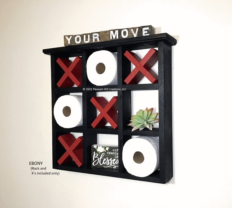May include: A black wooden wall-mounted shelf with a tic-tac-toe design. The shelf holds toilet paper rolls, red X's, and white O's. The top of the shelf has the words "YOUR MOVE" in white letters. A small sign reads "our family is blessed".