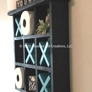 May include: Ebony-colored, wall-mounted shelf with nine compartments, designed to resemble a tic-tac-toe board. The shelf features teal X's and O's, with the words "YOUR MOVE" on top. Includes two rolls of toilet paper and a small decorative plant.