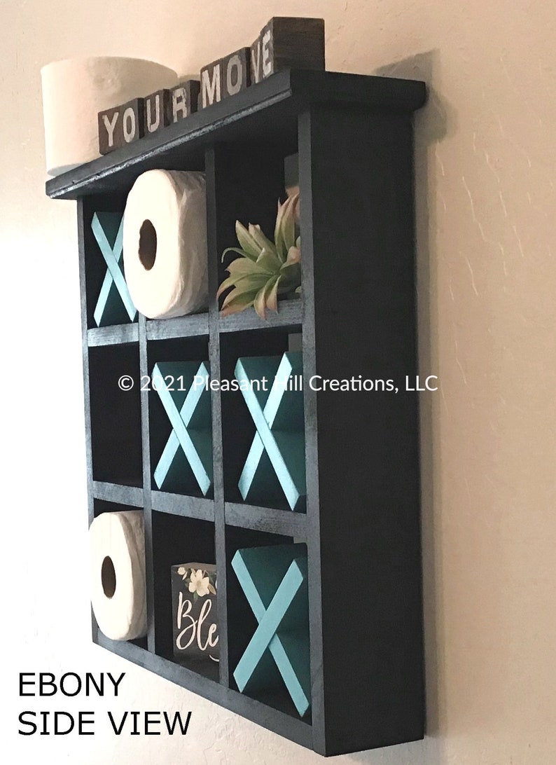 May include: Ebony-colored, wall-mounted shelf with nine compartments, designed to resemble a tic-tac-toe board. The shelf features teal X's and O's, with the words "YOUR MOVE" on top. Includes two rolls of toilet paper and a small decorative plant.