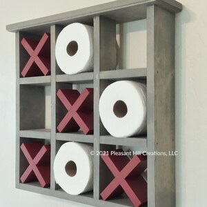 May include: A gray wooden wall-mounted organizer with nine compartments. The organizer holds three rolls of white toilet paper and six red X's. The text "SUNWASHED GRAY" is printed at the bottom. The organizer is designed to look like a tic-tac-toe board.