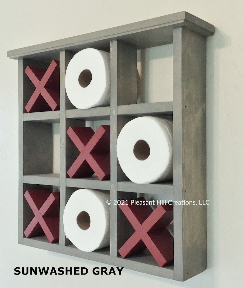 May include: A gray wooden wall-mounted organizer with nine compartments. The organizer holds three rolls of white toilet paper and six red X's. The text "SUNWASHED GRAY" is printed at the bottom. The organizer is designed to look like a tic-tac-toe board.