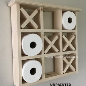May include: Unpainted wooden tic-tac-toe toilet paper holder with three rolls of white toilet paper. The holder has a grid design with X and O shapes. The text "UNPAINTED" is visible at the bottom.