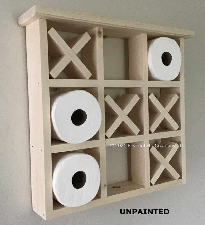 May include: Unpainted wooden tic-tac-toe toilet paper holder with three rolls of white toilet paper. The holder has a grid design with X and O shapes. The text "UNPAINTED" is visible at the bottom.