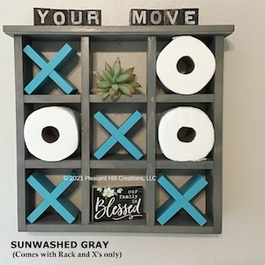 May include: A gray wooden tic-tac-toe game with turquoise X's and spaces for toilet paper rolls. The top has the words "YOUR MOVE" in block letters. A small sign reads "our family is Blessed".
