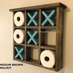 May include: A medium brown walnut tic-tac-toe-themed toilet paper holder. The wooden organizer features nine compartments, with three holding white toilet paper rolls and the others displaying teal X's and O's. The text "MEDIUM BROWN WALNUT" is at the bottom.