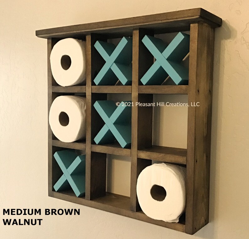 May include: A medium brown walnut tic-tac-toe-themed toilet paper holder. The wooden organizer features nine compartments, with three holding white toilet paper rolls and the others displaying teal X's and O's. The text "MEDIUM BROWN WALNUT" is at the bottom.
