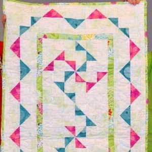 May include: A white quilt with a green, pink, and blue geometric pattern. The quilt has a border of green and white fabric. The quilt is labeled "quiltingjeanie designs".