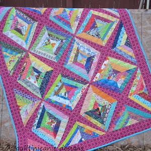 May include: A colorful quilt with a magenta background and a light blue border. The quilt features a patchwork design with various geometric shapes and patterns in shades of blue, green, orange, and pink. The text "quiltinjeanie designs" is visible.