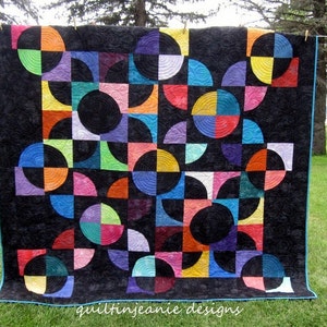 May include: A colorful quilt with a geometric pattern of circles and semi-circles on a black background. The quilt is hanging on a clothesline outside. The quilt is made of various fabrics in shades of red, orange, yellow, green, blue, purple, pink, and black. The quilt is quilted with a swirling pattern. The text "quiltingjeanie designs" is visible at the bottom of the quilt.