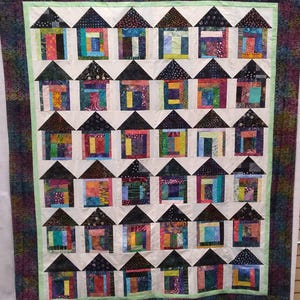 Pode incluir: Um edredom colorido com fileiras de casas com telhados pretos e paredes de patchwork vibrantes. As casas são dispostas sobre um fundo branco, emolduradas por uma borda verde e uma borda externa estampada. O edredom apresenta uma variedade de tecidos e cores.