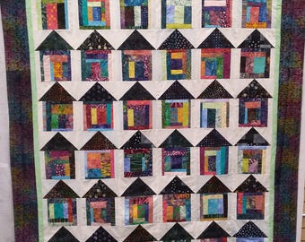 May include: A patchwork quilt with a repeating pattern of houses. Each house has a black roof and walls made from a variety of colourful fabrics. The houses are set against a white background, with a green border and a patterned outer edge.