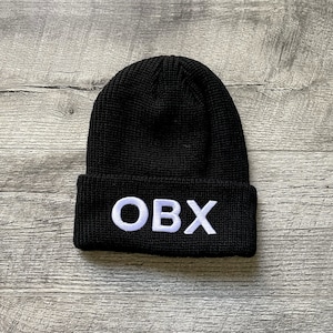 May include: Black knit beanie with the white embroidered letters "OBX" across the front. The hat is made of a textured material and is folded at the brim. The beanie is displayed on a wooden surface.