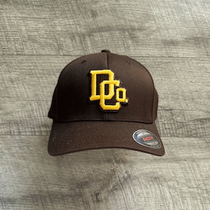 Può includere: Un cappello da baseball marrone con un logo ricamato giallo che dice "D.Co."