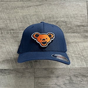 Op de afbeelding: Een donkerblauwe baseballpet met een oranje en witte lachende beer-patch op de voorkant.