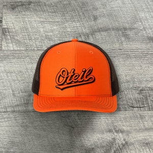 May include: Orange and black trucker hat with the word "Oteil" embroidered in black on the front.