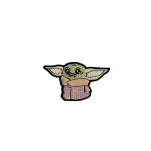 Puede incluir: Parche bordado con un personaje alienígena verde con orejas grandes y ojos grandes. El personaje lleva una túnica marrón y tiene una expresión neutra. El parche está delineado en negro sobre un fondo blanco.