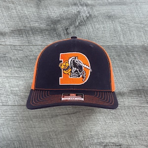 May include: A navy blue and orange baseball cap with a white embroidered logo of a bear and a lightning bolt. The logo is on an orange background and says "D". The cap is a trucker style with a mesh back.