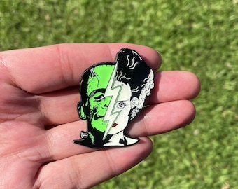 May include: A black and white enamel pin featuring a split image of Frankenstein and his bride. The image is divided by a lightning bolt. The pin is a green and white color.