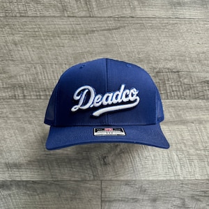 Może przedstawiać: Czapka trucker w kolorze królewskiego błękitu z haftowanym białym logo z napisem "Deadco" w stylu ozdobnym.