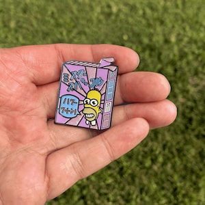 Puede incluir: Un pin rectangular de esmalte con una imagen de dibujos animados de Homer Simpson en una caja de cereales morada y rosa. El pin tiene texto japonés y una pequeña pestaña en la parte superior. El fondo es verde.