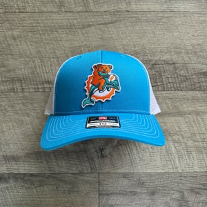 May include: A blue and white trucker hat with a white mesh back. The front of the hat features a patch of a cartoon bear riding a dolphin. The patch is in teal, orange, and white. The hat has a black adjustable strap with the text "Richardson" and "112" printed on it.