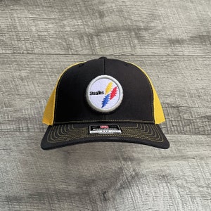 Pode incluir: Boné de caminhoneiro preto e amarelo com um patch circular com a palavra "Steelers" e um design de raio em vermelho, amarelo, azul e branco.