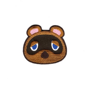 May include: Embroidered patch featuring the face of Tom Nook from the Animal Crossing video game series. The patch is brown, with a black nose, blue eyes, and pink ears.