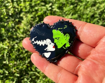 May include: Black and green heart-shaped pin with a black and white image of Frankenstein's monster and his bride kissing.