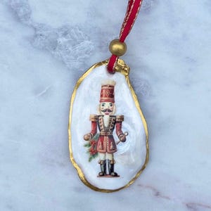 May include: A Christmas ornament with a nutcracker soldier design on a white oyster shell with gold trim. It hangs from a red ribbon and gold bead. The nutcracker wears a red uniform and black boots. The shell is approximately 8 cm long.
