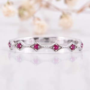May include: A silver band ring featuring a series of small, round, vibrant red rubies set within individual diamond-shaped metal frames. The metal surface has a textured, milgrain-style edge detail surrounding each of the inset red gemstones.