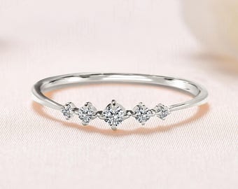 May include: A delicate silver ring featuring five small, sparkling diamonds set in a row. The ring has a thin band and is displayed on a soft, pink background, showcasing its elegant design. The diamonds are clear and catch the light.