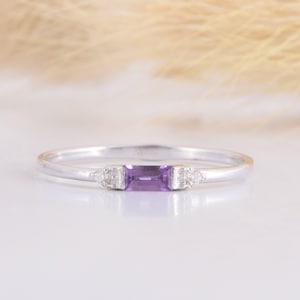 May include: A delicate silver ring with a rectangular purple amethyst gemstone and two smaller clear gemstones on either side.