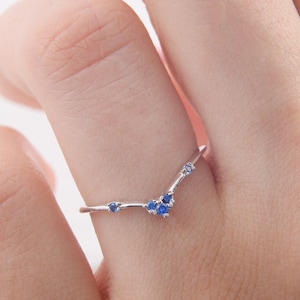 Op de afbeelding: Een zilveren ring met een V-vormig ontwerp bezet met kleine blauwe edelstenen.