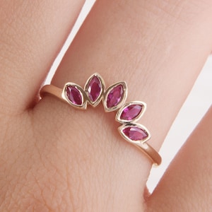 May include: A gold ring with five marquise-shaped ruby stones set in a curved band. The ring is worn on a finger.