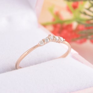 May include: A delicate rose gold band ring with a row of small, clear diamonds set in a curved, V-shaped design.