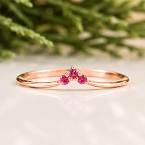 May include: A rose gold V-shaped ring featuring three small, round, vibrant pink gemstones set in a cluster at the centre point of the band. The metal band has a smooth, polished finish and a delicate, thin profile that curves into a chevron shape.