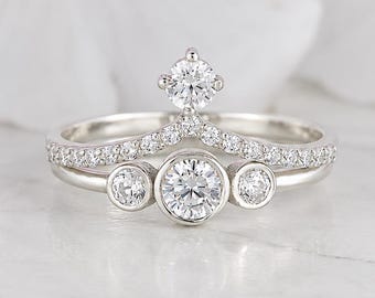 May include: A silver-toned ring set consisting of two stacked bands. The bottom band features three round, bezel-set stones. The top band is V-shaped with a row of small pavé-set stones and a single round stone centered at the peak of the V.