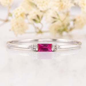May include: A thin silver-toned ring featuring a central rectangular baguette-cut vibrant pink gemstone. The stone is flanked on each side by three small, clear, round-cut accent stones set into the band, creating a delicate and refined appearance.