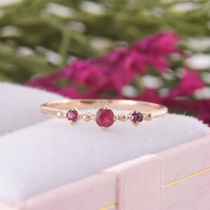 May include: A rose gold ring featuring three round, deep pink rubies set in a row. The center ruby is slightly larger than the two flanking stones. The band is thin and polished, with small metal bead accents placed between each gemstone setting.
