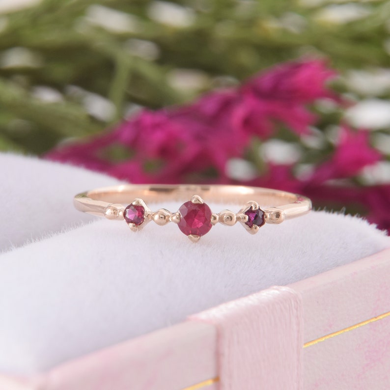May include: A rose gold ring featuring three round, deep pink rubies set in a row. The center ruby is slightly larger than the two flanking stones. The band is thin and polished, with small metal bead accents placed between each gemstone setting.