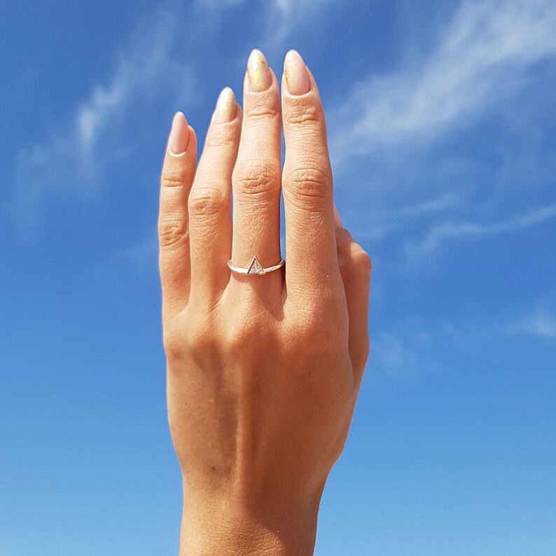 May include: A silver ring with a small triangle-shaped gemstone. The ring is on a person's finger against a blue sky background.