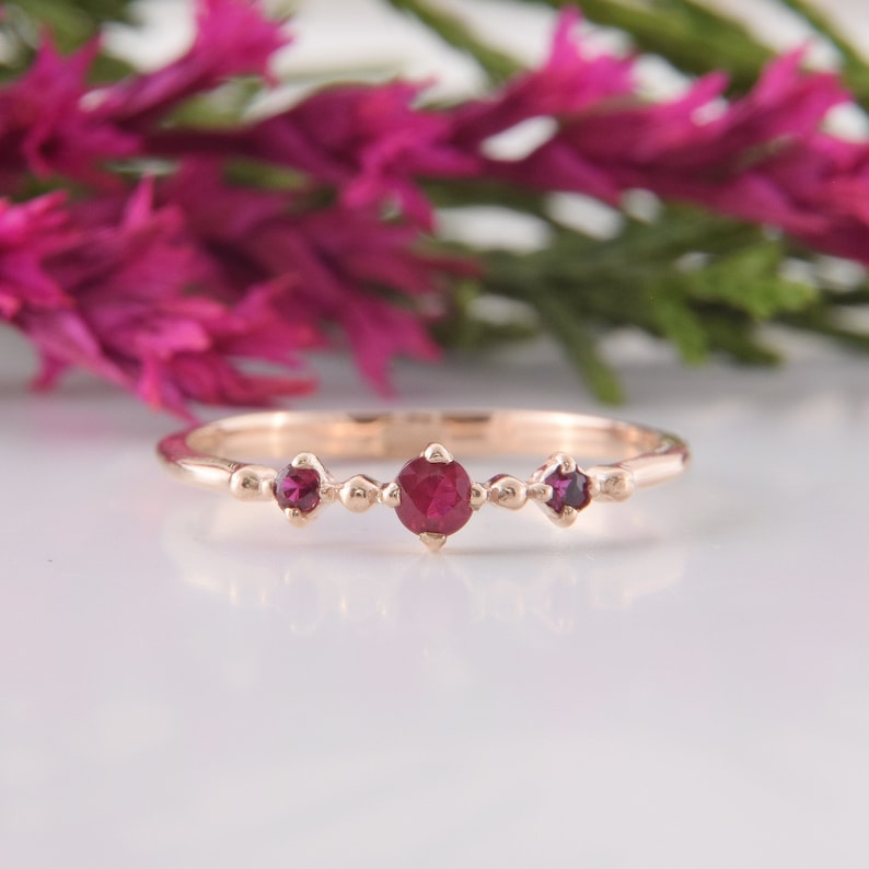 May include: A rose gold ring featuring three round, deep red rubies set in a row. The center ruby is slightly larger than the two smaller rubies on either side. The band is thin and smooth, with small metal bead accents between the stones.