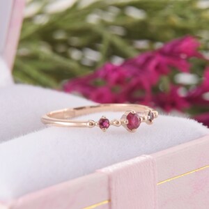 May include: A rose gold ring featuring three round, deep red rubies set in a row. The center ruby is the largest, flanked by two smaller rubies on either side. The band is thin and smooth, showcasing a delicate metal design with small bead accents.