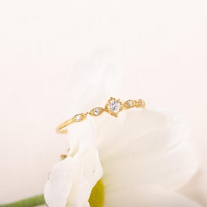 Pode incluir: Um delicado anel de ouro com um pequeno diamante redondo no centro e diamantes menores de cada lado. O anel est&aacute; colocado em uma flor branca.