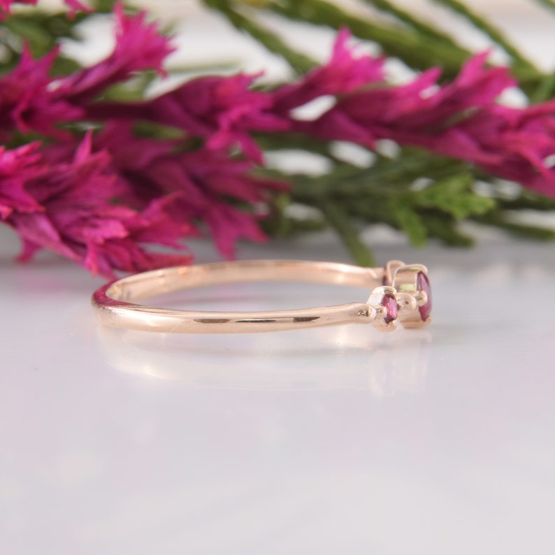 May include: A thin rose gold ring featuring two small, round, deep pink gemstones set side-by-side. The band is smooth and polished, with the stones held in place by small metal prongs. The ring is shown from a side profile angle.